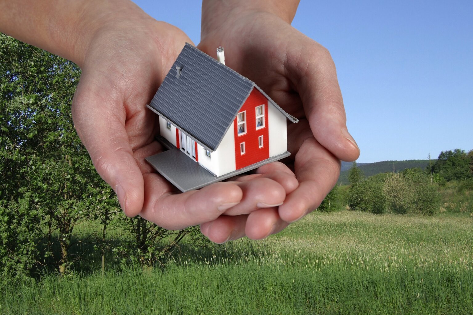 Hände halten ein kleines Einfamilienhaus-Modell über grüner Wiese – Symbolbild für unabhängige Begutachtung beim Hauskauf